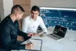 mentwo men sitting at a table in front of a laptop looking at business analytics